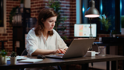 Fototapeta premium Office clerk looking through financial business documents on laptop late at night in workplace. Employee working on company project with deadline approaching soon during nightshift