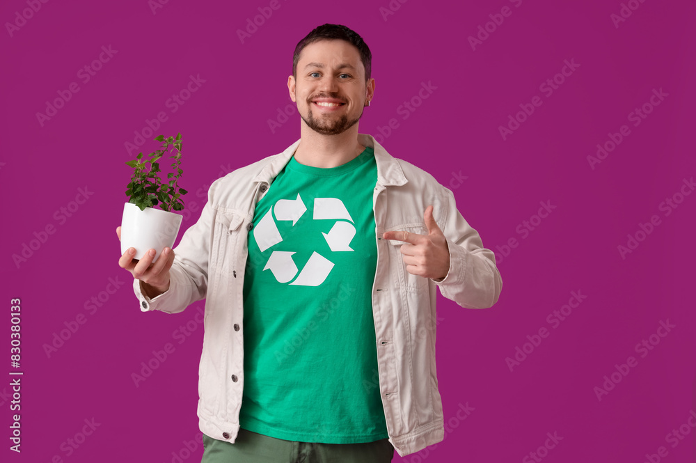 Wall mural happy young man in t-shirt with recycling logo pointing at houseplant on pink background. ecology co