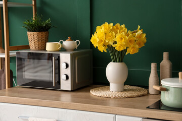 Interior of stylish kitchen with counter, microwave oven and vase of narcissus near green wall,...