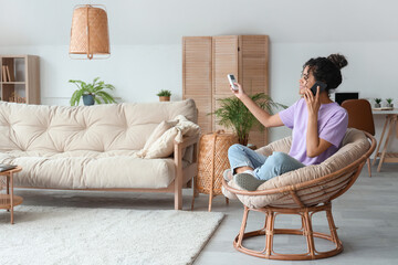 Young African-American woman with mobile phone in armchair turning on air conditioner at home