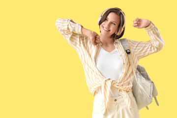 Happy female student with backpack and headphones dancing on yellow background