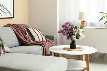 Beautiful living room with bouquet of lilacs branches on coffee table, cozy sofa and cushions