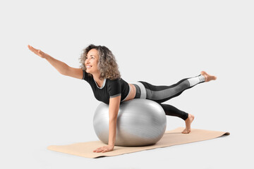 Sporty mature woman doing yoga on fitball against light background