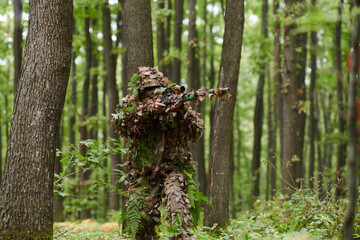A highly skilled elite sniper, camouflaged in the dense forest, stealthily maneuvers through...