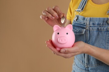 Woman putting coin into pink piggy bank on light brown background, closeup. Space for text