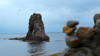 Stones on shore against background of cliff in sea. Clip. Stacked stones on rock background with flying seagulls in cloudy weather. Turrets of stones on shore at sea cliff with seagulls