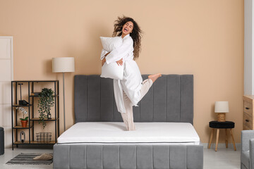 Young African-American woman with pillow jumping on soft mattress in bedroom