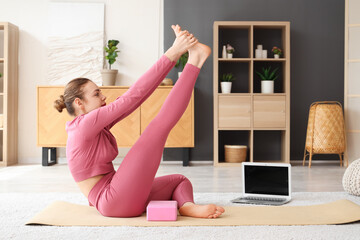 Sporty young woman stretching with yoga block and laptop on mat at home