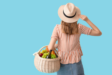 Beautiful young woman with food and bottle of wine for picnic in wicker basket on blue background, back view