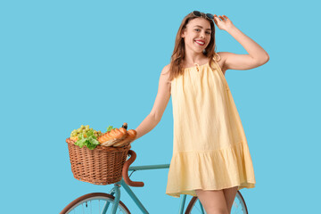 Beautiful young happy woman with bicycle and wicker picnic basket of tasty food on blue background