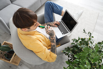 Young handsome man smoking disposable electronic cigarette and chatting with laptop on armchair at...