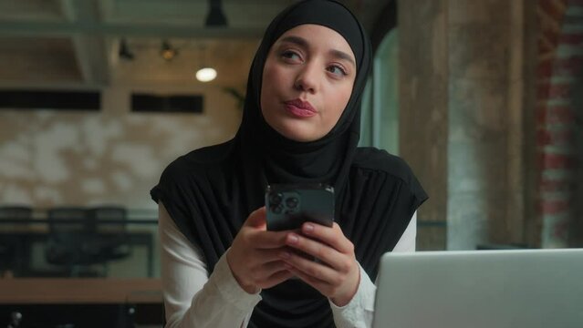Pensive thoughtful islamic muslim girl Arabian business woman in hijab female businesswoman office worker student think dream ponder consider idea using mobile phone thinking typing message smartphone