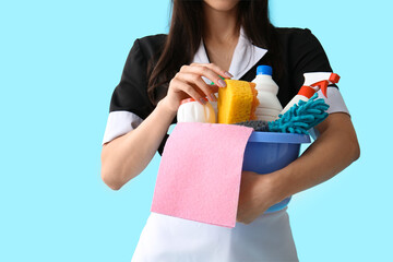 Young chambermaid with cleaning supplies on blue background, closeup