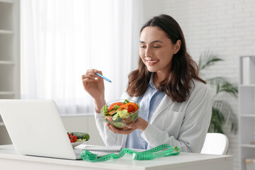 Young female nutritionist with vegetable salad video chatting at table in office