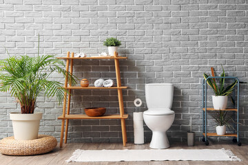 Interior of restroom with toilet bowl, plant and shelving unit near grey brick wall