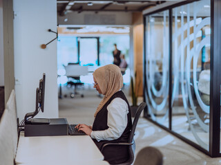 In a modern office, a young Muslim entrepreneur wearing a hijab sits confidently and diligently...