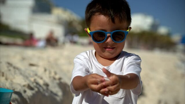 Slow motion of a cute young mexican latin boy wearing sunglasses and a white t-shirt showing some seashells to the camera and then putting them in a bucket