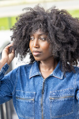 A confident woman with natural curls is wearing a denim jacket, exuding style and charm outdoors in...