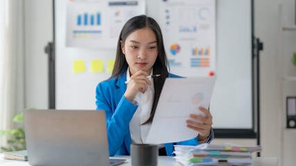 Businesswoman analyzing financial documents in modern office. Laptop and charts on whiteboard in...