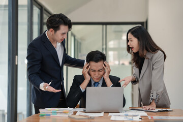 Stressed businessman surrounded by colleagues arguing while working on a laptop in a modern office setting, highlighting workplace tension.