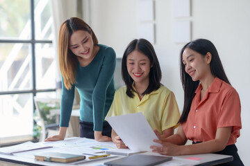 Three young businesswomen collaborating on a project while reviewing documents at a desk in a bright, modern office setting.