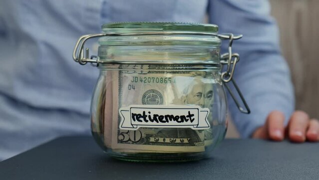 Unrecognizable woman put on table Glass jar full of American currency dollars cash banknote with text RETIREMENT. Preparation saving money. Moderate consumption and economy. Collecting money