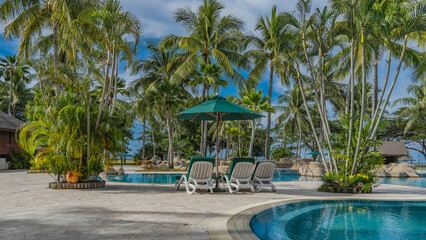 Deck chairs with soft mattresses and sun umbrellas are located by the swimming pool with clear turquoise water. Picturesque boulders along the perimeter. Tall coconut palms against a blue sky, clouds