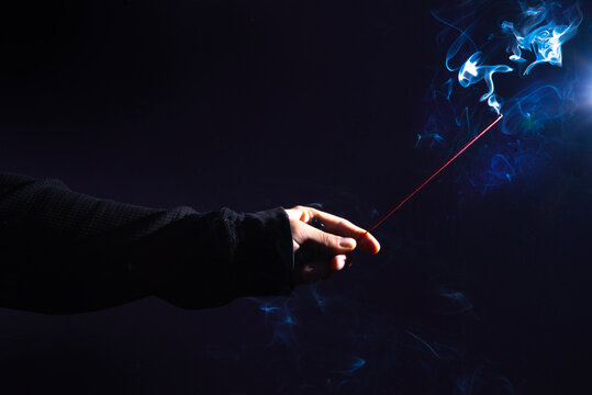 Mano, sosteniendo una varilla de incienso, en fondo negro con humo iluminado.