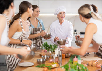 Elderly woman cook in white suit holds bowl in hands and tells women students of culinary courses...