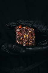 Hands in black gloves are holding chocolate bars mixed with nuts and fruits at black background