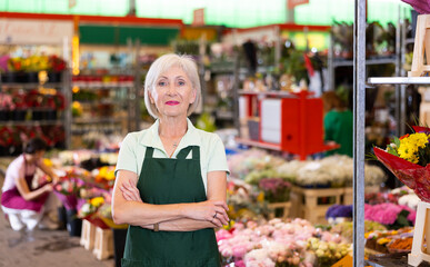 Portrait of successful experienced mature woman, floral shop owner, standing with crossed arms among stall with colorful blooming cut and potted flowers, looking at camera with smile
