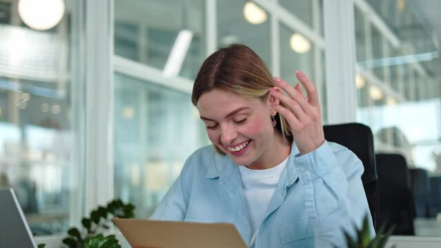 Cheerful successful female celebrating business achievements while holding envelope with pleasant news inside. Excited woman feeling happy because signing employment contract at personal workspace.