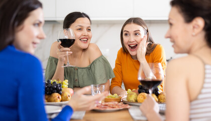 Cheerful female friends enjoying home gathering with wine and light snacks at kitchen table,...