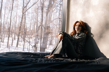 Portrait of pretty young redhead woman in casual clothes sitting resting by window with nature view on sunny winter day. Pensive attractive female sitting on floor. Feelings and emotions, free time