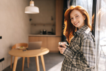 Cheerful young woman with red hair enjoying drinking cup of coffee at home standing by window on sunny morning, looking at camera with happy expression. Smiling pretty female drinking hot tea.