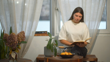 Young female student reads book in cafe. Media. Beautiful young woman comfortably spending time reading book in cafe. Cozy winter day in cafe with book and tea