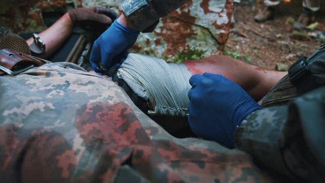 Close up hands combat medic with gloves fixes the leg with a tourniquet and bandage of wounded soldier. Blood around. Give first aid. Military concept. Training