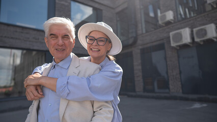 A woman in a hat and glasses hugs an elderly man in a white jacket from behind. Romantic relationships of mature people. 