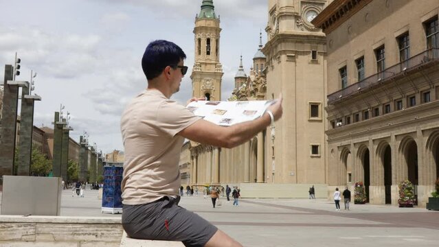 Young male tourist sitting in The Plaza of Our Lady of the Pillar in Zaragoza and looking at a paper map