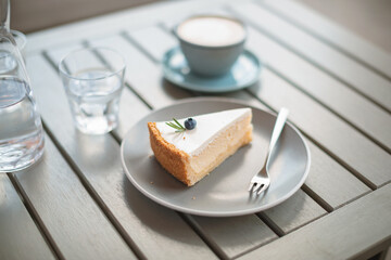 A piece of classic cheesecake decorated with a berry and rosemary on a plate, cup of coffee on the table on the terrace.
