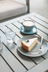 A piece of classic cheesecake decorated with a berry and rosemary on a plate, cup of coffee on the table on the terrace.