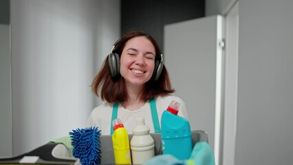 A cheerful brunette cleaning lady listens to music in black wireless headphones walks along the apartment corridor and carries a gray plastic basin with cleaning tools and detergents