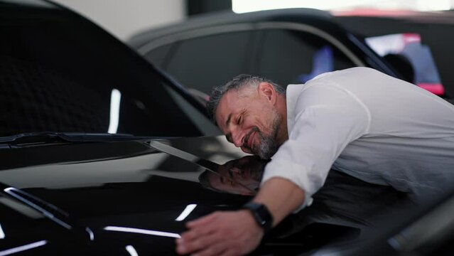 A happy middle-aged man with gray hair and strong stubble in a white shirt leans on a black car and hugs it happy And his purchases at a car dealership