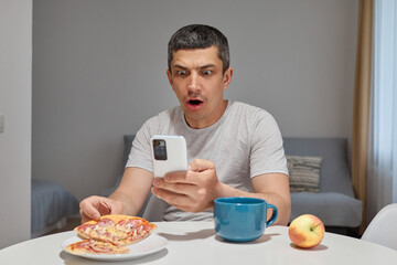 Shocked surprised astonished Caucasian man wearing T-shirt sitting at table in home interior holding smartphone reading internet news looking at device screen with big eyes