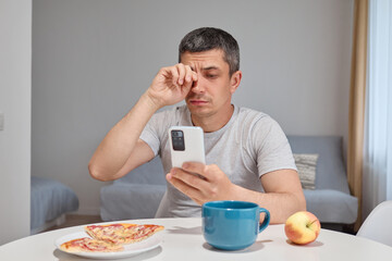 Tired exhausted Caucasian man wearing T-shirt sitting at table in home interior using mobile phone long hours rubbing his eyes feeling fatigue while browsing web pages
