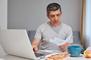 Serious concentrated Caucasian man wearing gray T-shirt working on laptop paying bills holding...