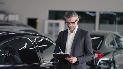 Portrait of a confident middle-aged man manager at a car dealership who stands in a white suit with a tablet in his hands and wearing glasses in the center of a car dealership