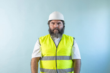 stressed adult man engineer with beard and wearing helmet and safety vest, looking at camera on...