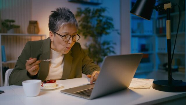 Mature businesswoman works till late night on laptop, eats cake, unbalanced diet