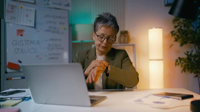Businesswoman works on laptop till late, checks her watch, feeling fatigue
